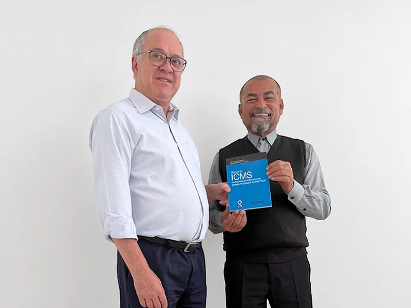 Os fundadores Marco Aurélio Guimarães Pereira e Alcides Joaquim Bonfim, de pé no escritório; Alcides segura o Manual do ICMS no Transporte Rodoviário de Cargas, de capa azul.