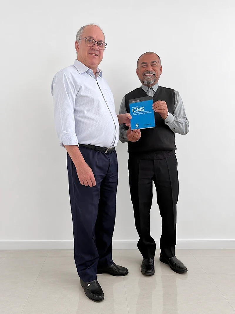 Marco Aurélio Guimarães Pereira e Alcides Joaquim Bonfim, fundadores da Paulicon, de pé no escritório; Alcides segura o Manual do ICMS de capa azul.